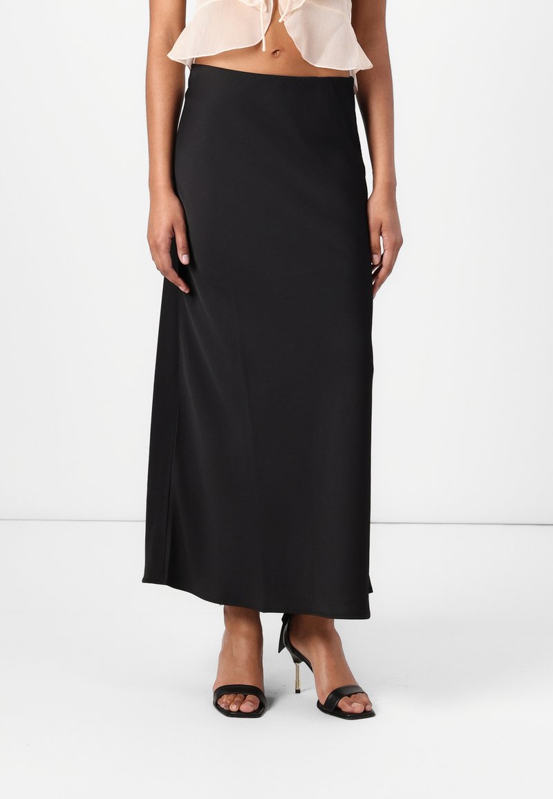 Jupe maxi en satin noir avec une texture lisse, dotée d'une taille ajustée et d'une légère fente sur le côté. Portée avec des sandales à talons aiguilles noires à brides.