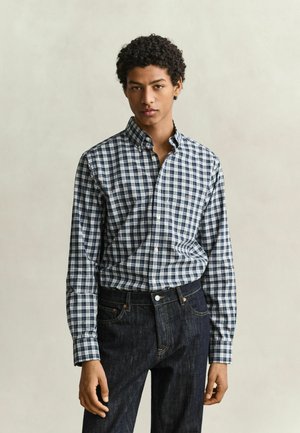 Jonge man met krullend haar, die een blauw-wit geruit overhemd met knopen en donkerblauwe spijkerbroek draagt tegen een effen achtergrond.