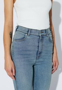 Mujer con jeans de tiro alto de color azul claro y una camiseta sin mangas de canalé blanca, de pie con los brazos relajados a los lados.