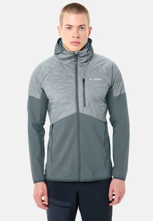 Young man wearing a gray two-tone zip-up jacket with a hood, standing straight against a plain light background.