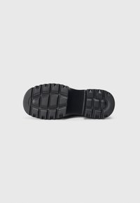 Semelle de chaussure en caoutchouc noir avec un motif texturé et robuste, présentant des rainures profondes et des sections surélevées pour une traction améliorée.