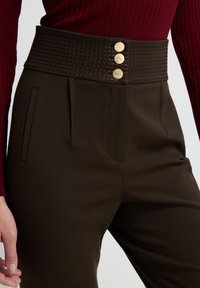 Pantalones marrones con un cinturón texturizado de talle alto que presenta tres botones en tono dorado, bolsillos laterales y detalles plisados.