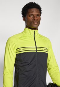 Leichte Jacke mit hohem Kragen, die über leuchtend gelbe und dunkelgraue Einsätze, horizontale Streifen und einen Reißverschluss verfügt. Markenlogo sichtbar.