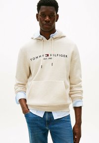 Felpa crema con cordini e tasca frontale, con logo "TOMMY HILFIGER EST. 1985". Maniche a camicia a righe blu visibili sotto. Jeans in denim.