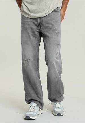 Mann trägt lockere, graue, abgenutzte Jeans und grauweiße Turnschuhe, steht mit einer Hand in der Tasche vor einfachem Hintergrund.