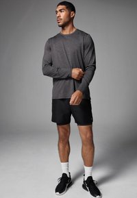 Långärmad, mörkgrå sporttröja med subtila ränder, tillsammans med svarta shorts och svarta sportskor. Modellen står framför en neutral bakgrund.