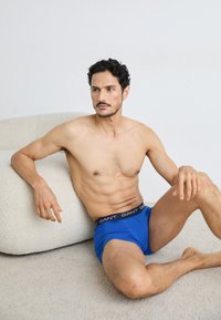 Modré boxershorts vyrobené z bavlny, s čiernym pásom so značkou "GANT". Model má štíhlu postavu a sedí na svetlej ploche.