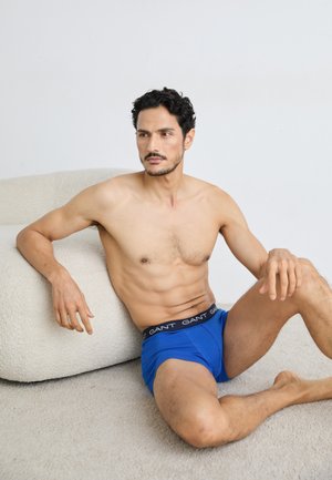 Modré boxerky z bavlny s černým páskem, na kterém je logo "GANT". Model má štíhlou postavu a sedí na světlém povrchu.