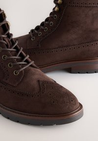 Hnedé semišové čižmy s brogue detailmi, okrúhla špička, kovové očká a tmavá gumová podrážka. Šnúrky sú stredne hnedé a dopĺňajú semiš.