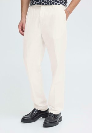 Lichtbeige broek met rechte pasvorm, voorzien van een platte voorkant en zijzakken. Draagt zwart gepolijst schoeisel. Gladde stoftextuur.