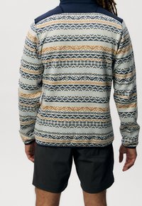 Veste en polaire avec motif beige, bleu marine et orange. Dotée de manches longues et d'un col montant, associée à un short foncé.