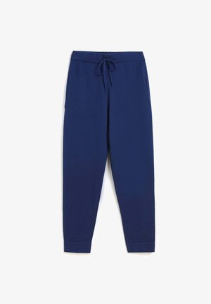 Person trägt dunkelblaue Jogginghose mit Kordelzug und weiße Sneakers, hebt einen hellblauen Pullover hoch, um einen definierten Bauch zu zeigen.