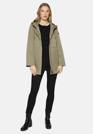 Kobieta ubrana w oliwkową kurtkę z kapturem, czarną górę z guzikami, czarne legginsy i czarne botki na obcasie, stojąca na białym tle.