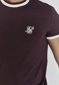 Bordeaux t-shirt met korte witte ribkraag en -manchetten. Bevat een wit geborduurd logo op de linkerborst. Gladde katoen structuur.