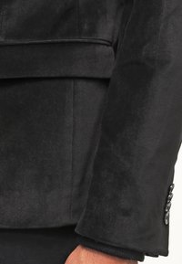 Rękaw czarnej aksamitnej marynarki o gładkiej fakturze, z klapami w kształcie litery V oraz detalem w postaci mankietów z trzema guzikami. Dopasowany krój z wyraźnymi szwami.