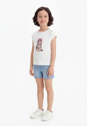 Lapsi seisoo valkoisessa t-paidassa, jossa on kissanpentu piirroksena vaaleanpunaiset aurinkolasit päässä, vaaleansiniset farkkushortsit ja valkoiset tennarit valkoisella taustalla.