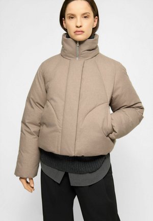 Kvinde iført en beige polstret jakke med høj hals med sorte bukser og lag-på-lag mørkegrå sweatere, stående mod en ensfarvet hvid baggrund.