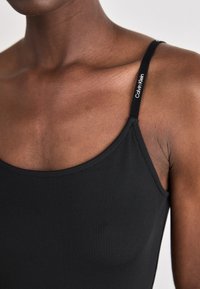 Czarny prążkowany top z cienkimi ramiączkami, z małym białym logo "Calvin Klein" na ramiączku. Materiał ma gładką fakturę.