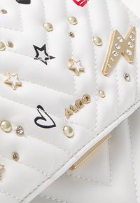 Valkoinen tikattu käsilaukku, jossa metalliset niitit, mustia ja punaisia koriste-elementtejä sekä kultainen logokoriste. Sileä pinta ja jämäkkä muoto.