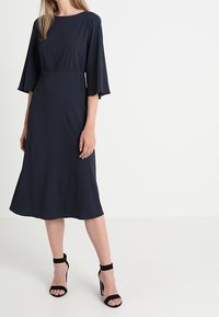 Žena v námořnickém midi šatu s rukávy ve tvaru zvonu po lokty a černými sandály na vysokém podpatku s kotníkovým páskem, stojící na bílém pozadí.