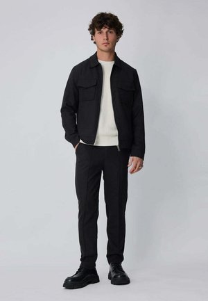 Jeune homme debout, portant une veste et un pantalon noirs, un pull blanc et des chaussures noires, sur un fond uni gris clair.