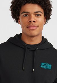 Schwarzer Hoodie mit einer Vorderasche, Kapuze mit Kordelzug und einem türkisfarbenen O'Neill-Logo-Patch auf der linken Brust. Weicher Stoff, lässiges Design.