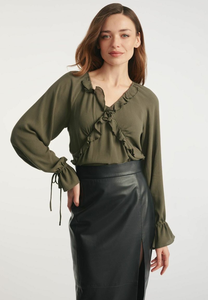 Oliwkowa bluzka z falbanami, długimi rękawami i wiązaniami, w zestawie z czarną skórzaną spódnicą ołówkową z bocznym rozcięciem.