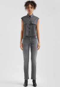 Camisa sin mangas de botón gris con dos bolsillos en el pecho, combinada con jeans ajustados grises y botas negras hasta el tobillo. Tejido suave y diseño ajustado.