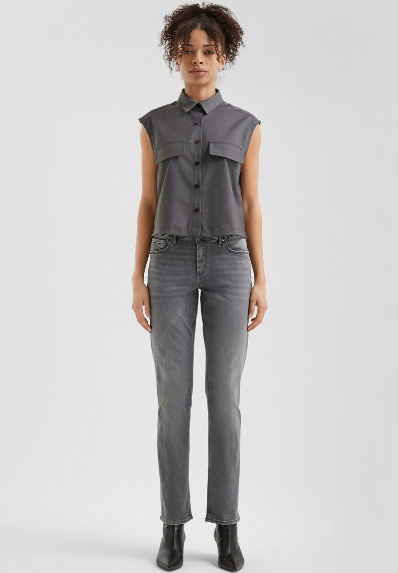 Camisa sin mangas de botón gris con dos bolsillos en el pecho, combinada con jeans ajustados grises y botas negras hasta el tobillo. Tejido suave y diseño ajustado.
