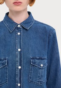 Τζιν πουκάμισο σε μεσαίο μπλε χρώμα, με γιακά, κουμπιά snap και διπλές τσέπες στο στήθος με καπίστρια. Απαλή υφή, χαλαρός σχεδιασμός.