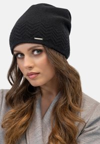 Berretto nero in maglia con motivo a zigzag, dotato di un'etichetta accentuata in argento. Il tessuto appare lavorato a maglia e accogliente, aderente alla testa.