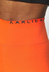 Korkeavyötäröiset, venyvät oranssit legginsit, joissa on musta logovyötärö. Sileä rakenne ja istuva muotoilu, joka sopii aktiiviseen pukeutumiseen.