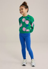 Zöld pulóver rózsaszín gyümölcs mintával, kék bordázott leggings, és ezüst sportcipők. Az öltözet élénk színekkel és kényelmes szabással rendelkezik.
