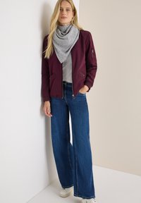 Kvinde med langt blondt hår iført et gråt tørklæde, maroon bomberjakke, store blå jeans og hvide sneakers, der står mod en hjørnevæg.