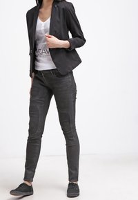 Schwarzes Blazer mit glatter Textur, kombiniert mit schlanken schwarzen Jeans mit Knietaschen, getragen mit einem weißen Grafik-T-Shirt und schwarzen Schuhen.