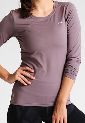 Kobieta ubrana w dopasowaną, długa, mauve sportową bluzkę z małym logo na piersi oraz czarne legginsy, pozująca z jedną ręką na biodrze.