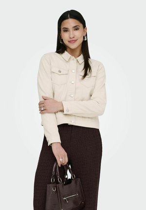 Ženska z dolgimi temnimi lasmi, oblečena v svetlo bež jakno in temne teksturirane hlače, drži temno rjavo torbico z obema rokama.