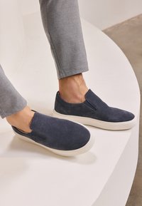 Marineblauwe suede instapschoenen met elastische zijkanten en een witte rubberen zool, gecombineerd met lichtgrijze tapered pants. Geweven oppervlak zichtbaar.