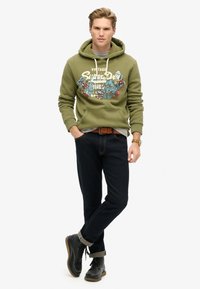 Oliwkowa bluza z kapturem z graficznym nadrukiem i sznurkiem, szary t-shirt pod spodem, ciemne dżinsy i czarne botki z podwiniętymi nogawkami.