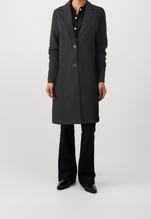 Kobieta ubrana w długi czarny płaszcz z dwoma guzikami, czarne spodnie i czarne buty w szpic, stojąca na tle jednolitego jasnego tła.