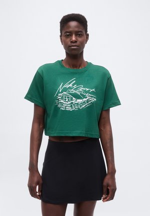 Personne portant un t-shirt court vert avec un graphisme et un texte blancs, associé à une jupe noire, debout devant un fond uni clair.