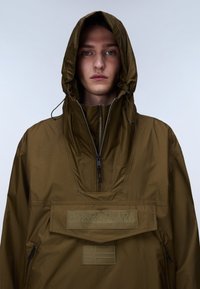 Ung person iført oliengrøn hættetrøje med lynlås foran og stort Napapijri-logo på brystet.