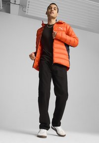 Pufferjakke i lys orange med sorte accenter, lynlåslukning og hætte, dansk med sorte bukser og hvide sneakers. Enkelt baggrund.