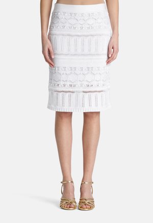 Jupe crayon blanche en maille avec des motifs texturés, taille côtelée, longueur genoux, associée à des talons dorés à brides avec des détails tressés.