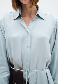 Cămașă din satin de culoare albastru deschis, cu guler, nasturi frontali și cordon la talie. Prezintă un model în gradient care trece la o nuanță de maro închis.
