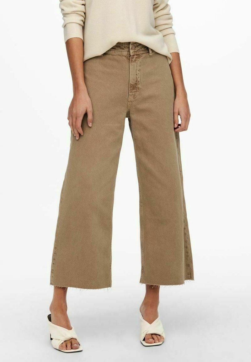 Jeans beige à jambes larges avec ourlet effiloché, fabriqués en denim robuste. Associés à des sandales crèmes nouées. Design simple et fonctionnel.