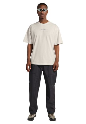 Mand iført hvid t-shirt med grafik, sorte cargobukser, chunky sneakers og hvide sporty solbriller, stående mod en hvid baggrund.