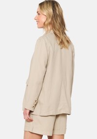Beige Leinenjacke mit lockerem Schnitt, Knopfakzenten an den Ärmeln und geradem Saum. Kombiniert mit passenden Shorts für einen stimmigen Look.