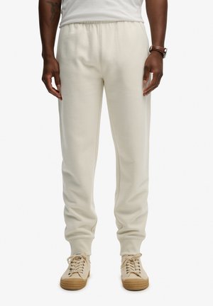 Homme portant un pantalon de survêtement écru et des baskets beiges à lacets, debout les mains détendues le long du corps.