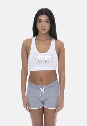 Femme aux longs cheveux foncés portant un soutien-gorge de sport blanc avec le texte "LEONE 1947" et un short gris avec cordon, debout devant un fond uni.